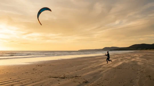 Un rider pratiquant un sport de traction sur une plage venteuse au coucher du soleil avec une aile colorée dans le ciel