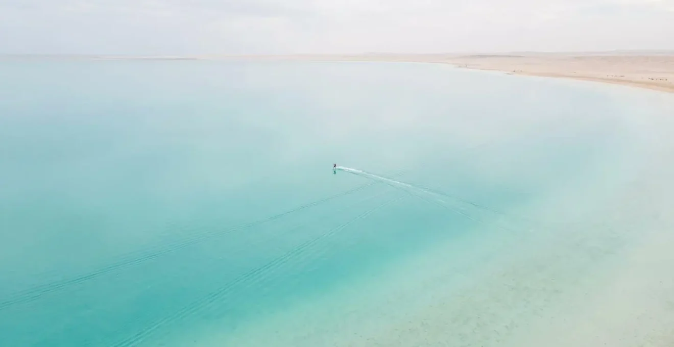 Vue aérienne minimaliste d'une vaste lagune turquoise aux eaux plates et peu profondes, un kitesurfeur solitaire créant des lignes symétriques à la surface